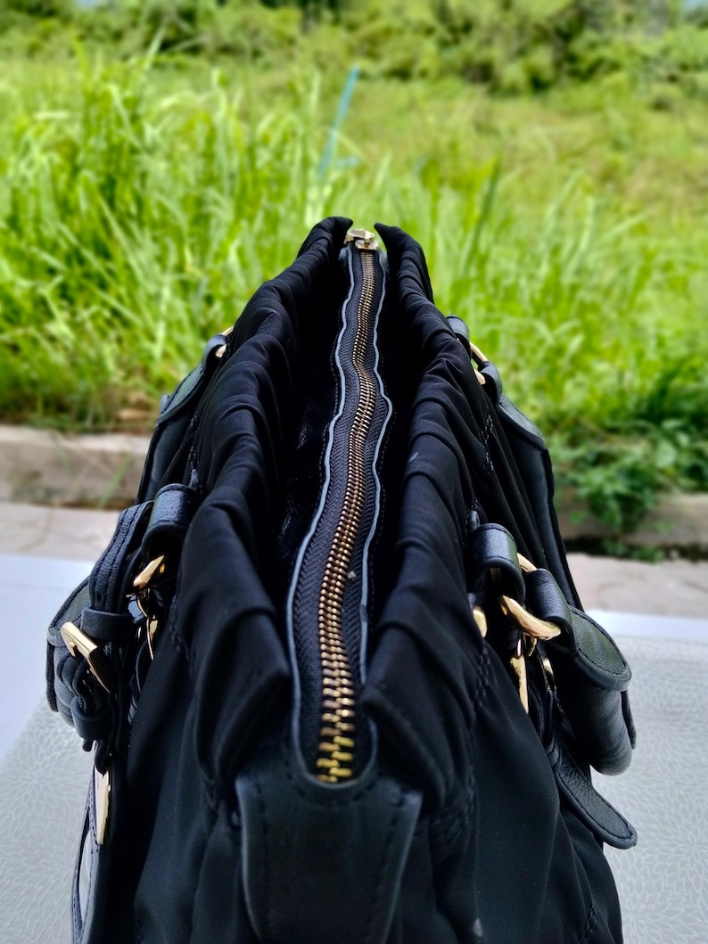 Puede incluir: Bolso de mano negro con cremallera y herrajes dorados. El bolso est&aacute; hecho de un material suave y fruncido, con detalles de cuero. La cremallera est&aacute; abierta, revelando el interior. El fondo es follaje verde borroso.