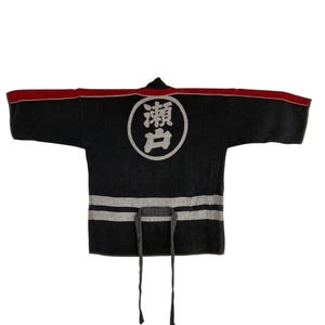 Puede incluir: Un abrigo de trabajo japonés gris oscuro con ribete rojo en los hombros. El abrigo presenta un emblema circular blanco con caracteres japoneses en la espalda y dos rayas horizontales blancas en la parte inferior. Tiene mangas largas y cierre de lazo.