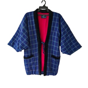 Puede incluir: Chaqueta estilo kimono a cuadros azules con detalles turquesa y negros. La chaqueta tiene un interior rojo y ribetes negros a lo largo de los bordes y bolsillos. Cuelga de una percha negra.
