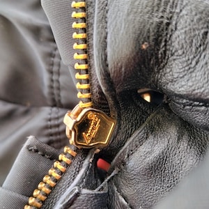 Puede incluir: Primer plano de una chaqueta negra con cremallera dorada. La cremallera est&aacute; completamente cerrada y el tirador dorado es visible. La chaqueta parece estar hecha de nailon o material similar, con textura de cuero o similar.
