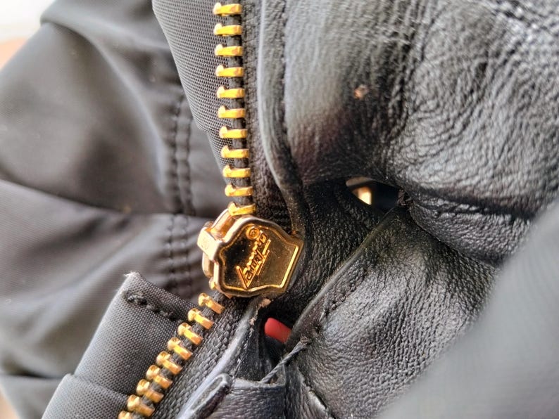 Puede incluir: Primer plano de una chaqueta negra con cremallera dorada. La cremallera est&aacute; completamente cerrada y el tirador dorado es visible. La chaqueta parece estar hecha de nailon o material similar, con textura de cuero o similar.