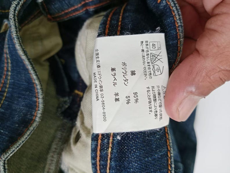 Puede incluir: Primer plano de una etiqueta de jeans de mezclilla con texto japon&eacute;s y s&iacute;mbolos de cuidado. La etiqueta indica que los jeans est&aacute;n hechos de 95% algod&oacute;n y 5% poliuretano. La etiqueta tambi&eacute;n indica "MADE IN CHINA".