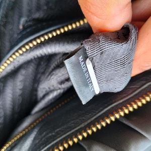 Puede incluir: Primer plano del interior de un bolso negro con cremallera dorada. Una etiqueta gris con el texto "MADE IN ITALY" es visible. El forro del bolso es de tela gris oscuro.