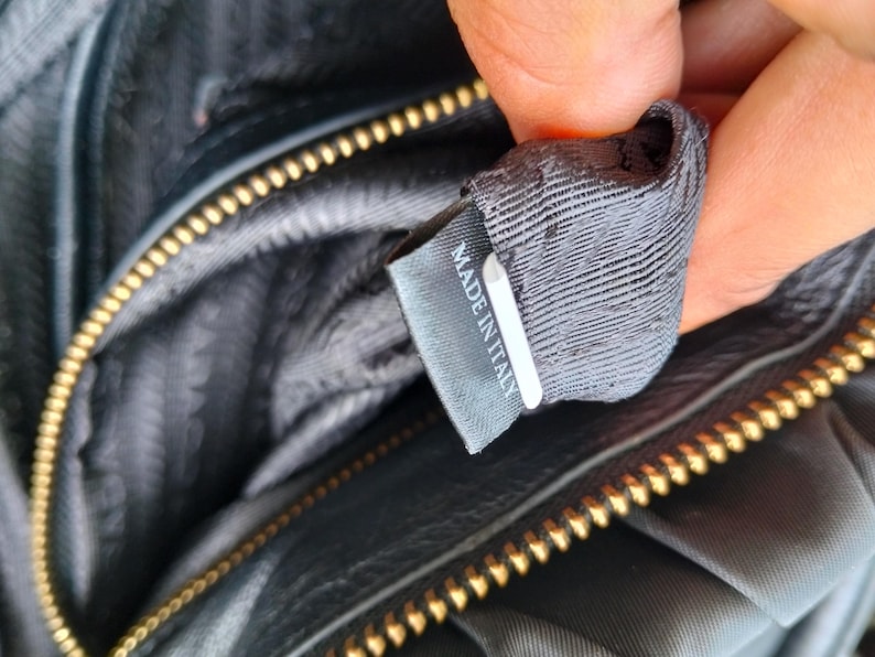 Puede incluir: Primer plano del interior de un bolso negro con cremallera dorada. Una etiqueta gris con el texto "MADE IN ITALY" es visible. El forro del bolso es de tela gris oscuro.