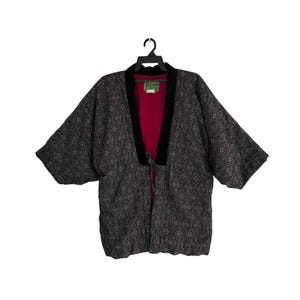 Op de afbeelding: Een kimono-achtige jas met patroon aan een zwarte hanger. De jas heeft een geometrisch ontwerp in grijstinten en zwart. De binnenvoering is dieprood en de kraag en bies zijn zwart.