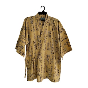 May include: A tan kimono-style robe with black calligraphy print. The robe has a V-neck, short sleeves, and a tie closure. The fabric has a subtle striped texture, and the robe is hanging on a black hanger.