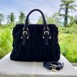 Puede incluir: Bolso de mano negro con asa superior y bandolera extra&iacute;ble. El bolso presenta herrajes dorados y hebillas decorativas. El bolso est&aacute; hecho de tela fruncida y se apoya sobre una superficie blanca.
