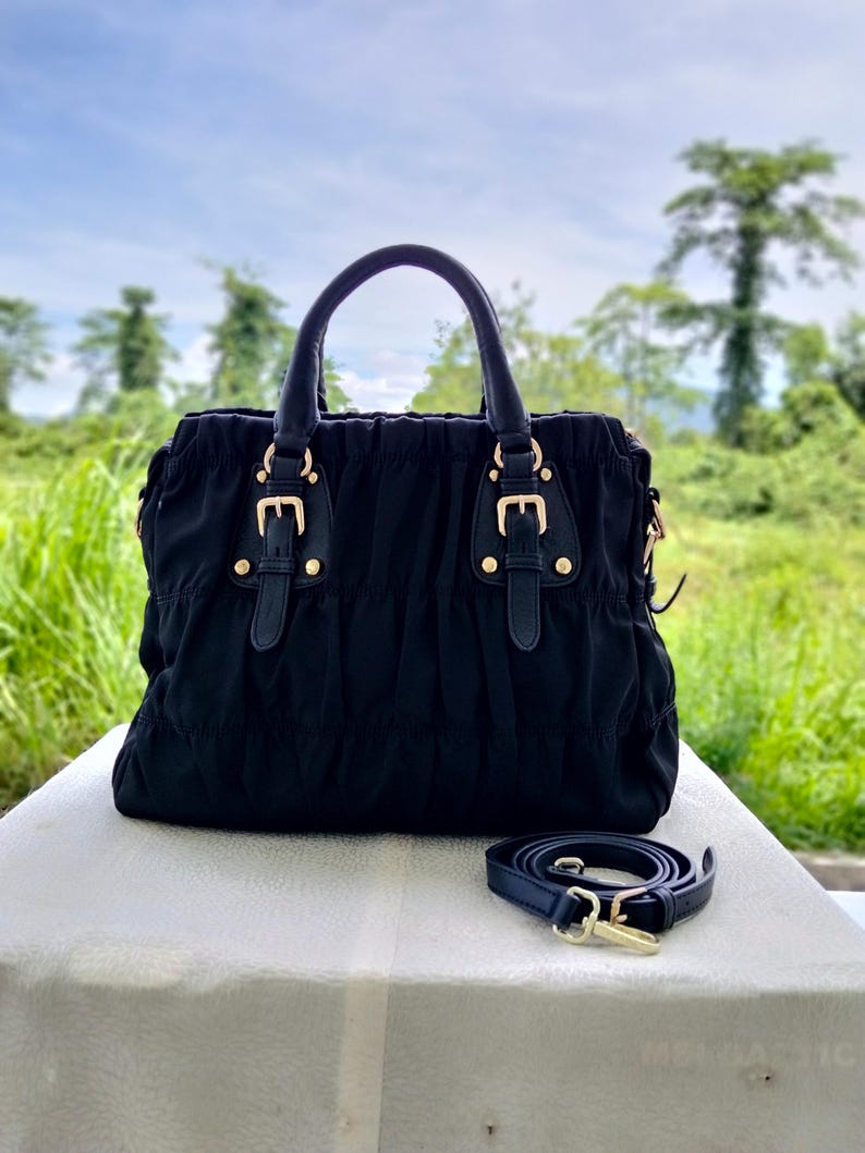 Puede incluir: Bolso de mano negro con asa superior y bandolera extra&iacute;ble. El bolso presenta herrajes dorados y hebillas decorativas. El bolso est&aacute; hecho de tela fruncida y se apoya sobre una superficie blanca.
