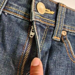 Puede incluir: Primer plano de unos vaqueros azules con costuras naranjas. Los vaqueros tienen una cremallera met&aacute;lica y un cierre de bot&oacute;n. El bot&oacute;n es de color cobre con un dise&ntilde;o detallado.