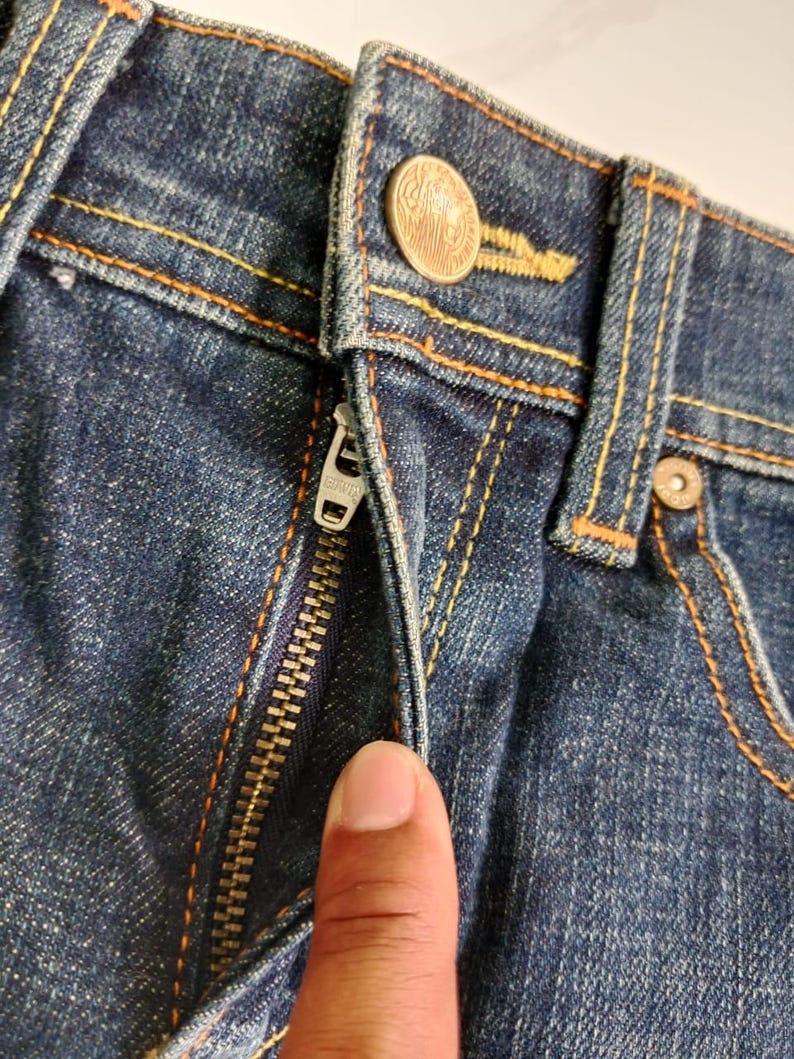 Puede incluir: Primer plano de unos vaqueros azules con costuras naranjas. Los vaqueros tienen una cremallera met&aacute;lica y un cierre de bot&oacute;n. El bot&oacute;n es de color cobre con un dise&ntilde;o detallado.