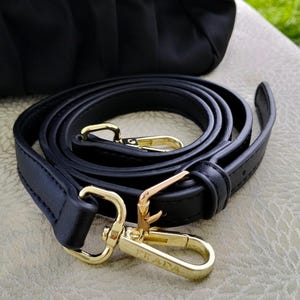 Puede incluir: Correa de hombro de cuero negro con herrajes dorados. La correa est&aacute; enrollada y presenta dos cierres dorados con el nombre de la marca "PRADA" grabado en uno de los cierres. Una bolsa negra es visible en el fondo.