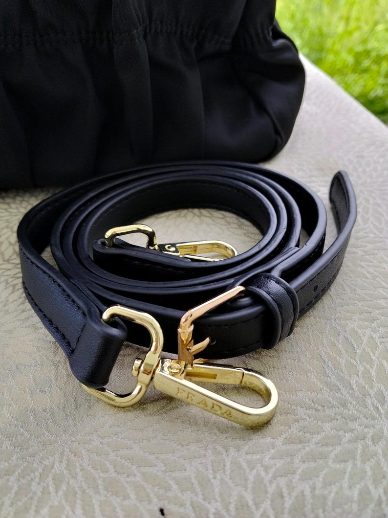 Puede incluir: Correa de hombro de cuero negro con herrajes dorados. La correa est&aacute; enrollada y presenta dos cierres dorados con el nombre de la marca "PRADA" grabado en uno de los cierres. Una bolsa negra es visible en el fondo.
