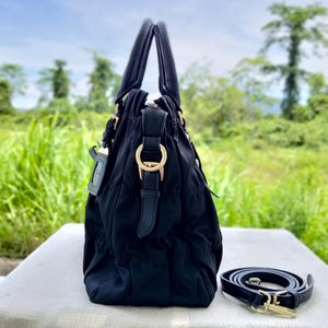Puede incluir: Bolso de mano negro con asa superior y correa para el hombro desmontable. El bolso presenta herrajes dorados y un dise&ntilde;o fruncido. El bolso est&aacute; sobre una superficie blanca con un fondo verde.