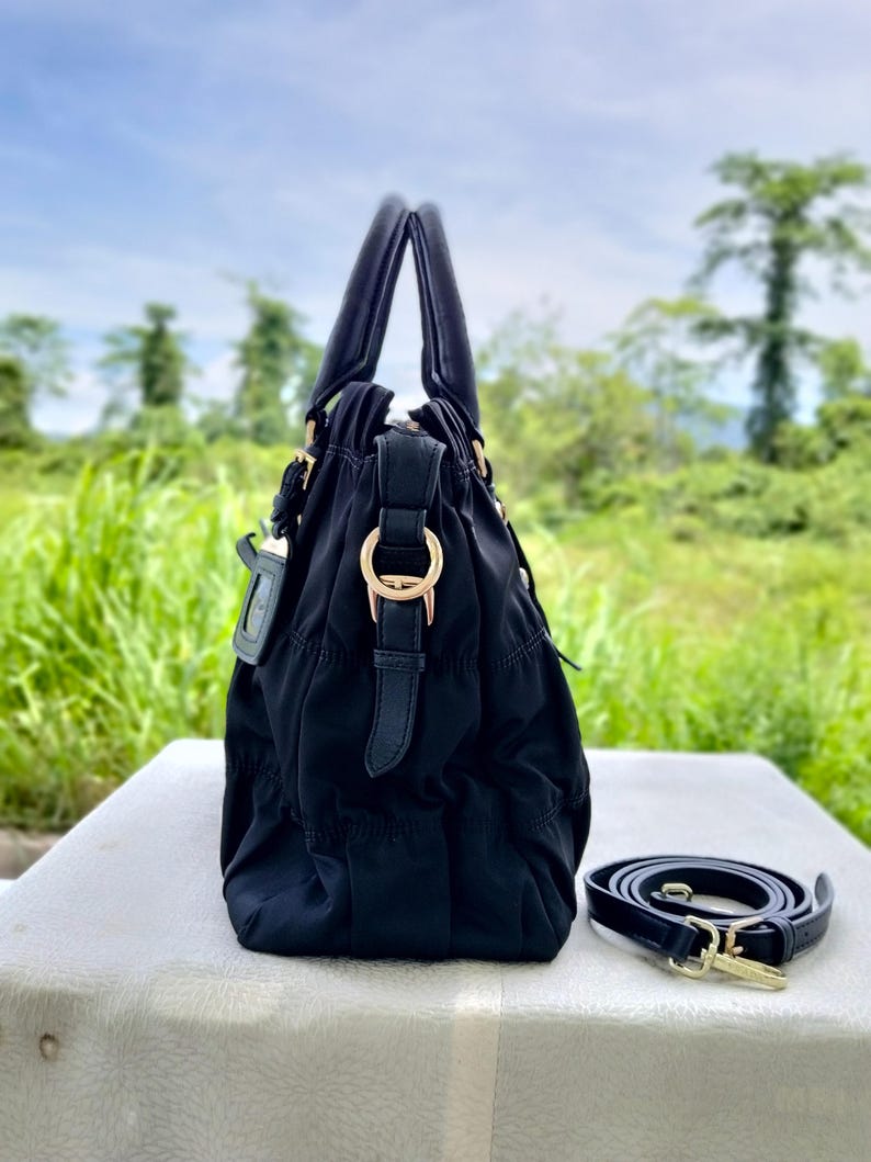 Puede incluir: Bolso de mano negro con asa superior y correa para el hombro desmontable. El bolso presenta herrajes dorados y un dise&ntilde;o fruncido. El bolso est&aacute; sobre una superficie blanca con un fondo verde.