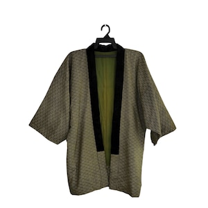 May include: Olive green kimono-style jacket with a geometric pattern. The jacket features black trim along the collar and front edges. The interior lining is a lighter shade of green. The garment is displayed on a black hanger.
