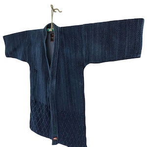 Puede incluir: Una chaqueta azul marino estilo kimono con un tejido texturizado. La chaqueta presenta un patrón de diamantes en la parte inferior y cuelga de un cordón de color natural. Las mangas son anchas y la chaqueta está abierta por delante.