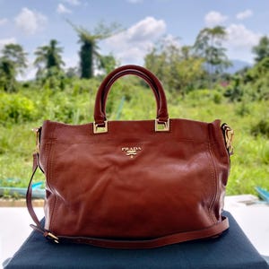 May include: A brown leather handbag featuring two top handles and a detachable shoulder strap. The front displays the gold-toned Prada logo hardware. The bag has a structured base with visible stitching details and gold-colored metal hardware accents.