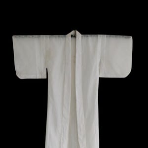 Puede incluir: Una bata blanca estilo kimono con mangas anchas, colgada sobre un fondo negro. La bata está hecha de una tela ligera y tiene un diseño simple y elegante. La bata está abierta por delante y parece sin usar.