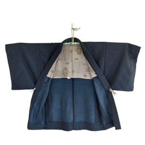 Könnte beinhalten: Ein dunkelmarineblauer Kimono mit weiten Ärmeln, der offen hängt, um ein gemustertes Innenfutter freizulegen. Der Kimono hängt an einem Holzbügel und zeigt sein traditionelles Design und seine Konstruktion. Das Innenfutter weist ein beiges Muster auf.