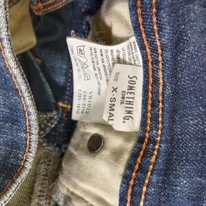 Puede incluir: Primer plano de unos vaqueros de mezclilla azules con costuras naranjas. Una etiqueta blanca dice "SOMETHING. EDWIN." y "X-SMALL." Los vaqueros tienen un bot&oacute;n y un interior beige. La tela es 95% algod&oacute;n y 5% poliuretano.