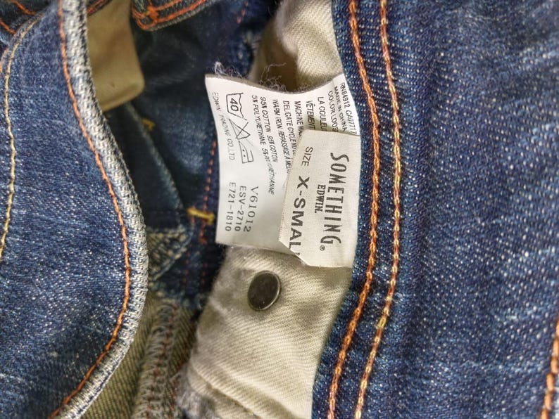 Puede incluir: Primer plano de unos vaqueros de mezclilla azules con costuras naranjas. Una etiqueta blanca dice "SOMETHING. EDWIN." y "X-SMALL." Los vaqueros tienen un bot&oacute;n y un interior beige. La tela es 95% algod&oacute;n y 5% poliuretano.