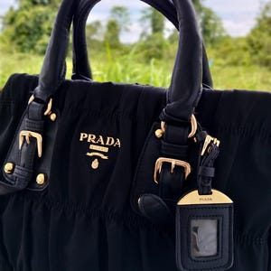 Puede incluir: Bolso Prada negro con herrajes dorados. El bolso presenta asas de cuero enrolladas, cierre de cremallera superior y una etiqueta de equipaje desmontable. El logotipo de Prada se muestra en la parte delantera.