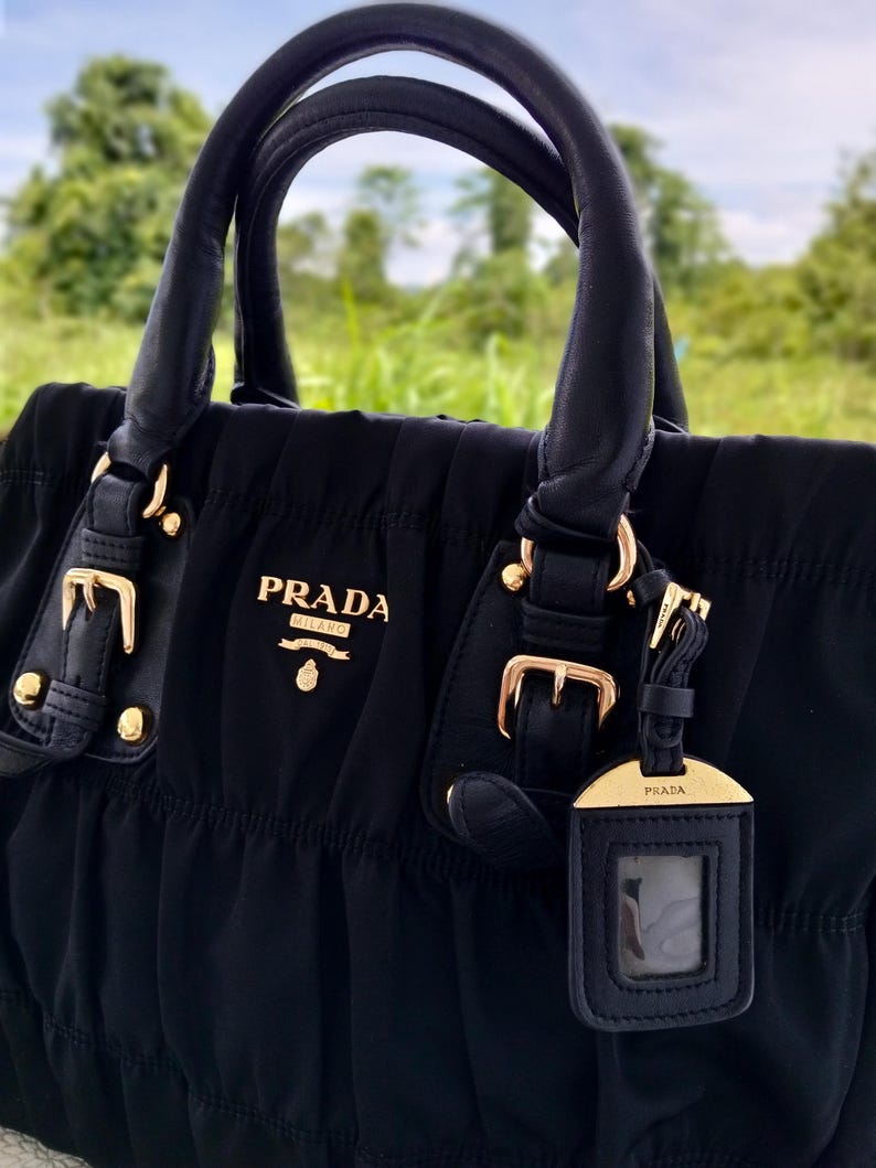 Puede incluir: Bolso Prada negro con herrajes dorados. El bolso presenta asas de cuero enrolladas, cierre de cremallera superior y una etiqueta de equipaje desmontable. El logotipo de Prada se muestra en la parte delantera.