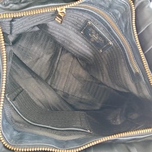 Puede incluir: Vista abierta de un bolso de mano de cuero negro con cremallera y herrajes dorados. El interior presenta un forro negro con un patr&oacute;n de logotipo repetido y un peque&ntilde;o bolsillo. Un logotipo dorado de Prada es visible dentro del bolso.