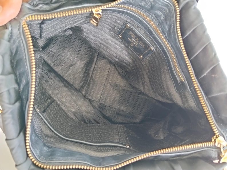 Puede incluir: Vista abierta de un bolso de mano de cuero negro con cremallera y herrajes dorados. El interior presenta un forro negro con un patr&oacute;n de logotipo repetido y un peque&ntilde;o bolsillo. Un logotipo dorado de Prada es visible dentro del bolso.