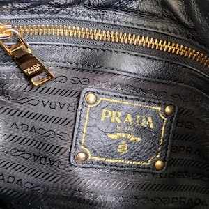Puede incluir: Primer plano del interior de un bolso de mano de cuero negro con cremallera dorada y logotipo Prada dorado. El logotipo Prada est&aacute; en un parche de cuero negro con detalles dorados.