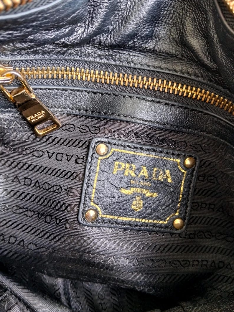 Puede incluir: Primer plano del interior de un bolso de mano de cuero negro con cremallera dorada y logotipo Prada dorado. El logotipo Prada est&aacute; en un parche de cuero negro con detalles dorados.