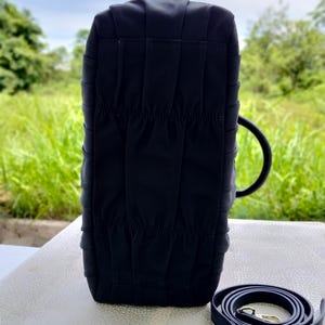 Puede incluir: Un bolso negro con un dise&ntilde;o estructurado, con una textura plisada y una correa para el hombro a juego. El bolso tiene un asa redondeada y herrajes dorados en la correa.