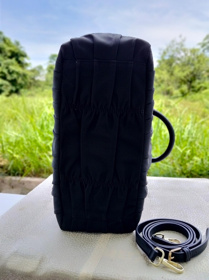 Puede incluir: Un bolso negro con un dise&ntilde;o estructurado, con una textura plisada y una correa para el hombro a juego. El bolso tiene un asa redondeada y herrajes dorados en la correa.