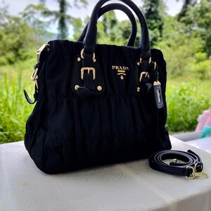 Puede incluir: Bolso de mano negro con herrajes dorados, con asa superior y correa para el hombro desmontable. El bolso tiene el nombre de la marca "PRADA" en letras doradas. El bolso est&aacute; hecho de una tela fruncida y tiene una forma estructurada.