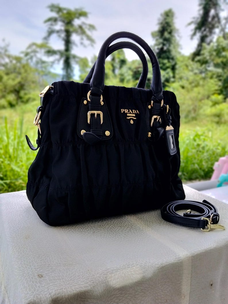 Puede incluir: Bolso de mano negro con herrajes dorados, con asa superior y correa para el hombro desmontable. El bolso tiene el nombre de la marca "PRADA" en letras doradas. El bolso est&aacute; hecho de una tela fruncida y tiene una forma estructurada.