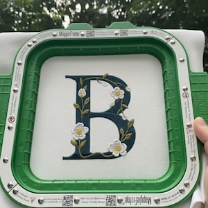 May include: An embroidered letter "B" in a dark teal color, adorned with white floral accents and green leaves, is displayed within a green embroidery hoop. The design is on a white fabric, showcasing a detailed floral monogram.