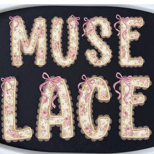 May include: The words "MUSE LACE" are presented in a decorative font. Each letter features a cream base with a floral pattern of pink roses and green leaves. The letters are outlined with gold trim and a scalloped lace edge, and adorned with pink bows.