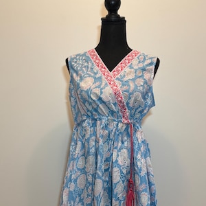 May include: A sleeveless blue wrap top with a floral pattern in white and pink. The top has a V-neckline with a pink trim and a matching pink tie with tassels. The garment is displayed on a black mannequin.