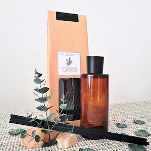 May include: An amber glass diffuser bottle with a black cap, black reeds, and a brown cardboard box. The box has the text "LUNA COLLECTIVE LONDON" and "O. BASILICUM". Decorative eucalyptus and small stones are in front of the bottle.