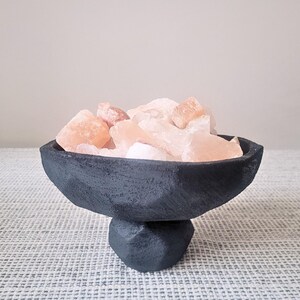 Puede incluir: Un cuenco con pedestal de color negro mate, formado a mano con una superficie rugosa y texturizada, lleno de rocas de sal del Himalaya translúcidas de color rosa pálido. El cuenco presenta una base ancha y poco profunda.