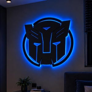 Peut inclure: Une enseigne murale noire représentant le logo des Transformers Autobot, composée d'un cadre circulaire et d'un masque stylisé. L'enseigne est éclairée par l'arrière par une lumière LED bleue vive, créant un effet de halo lumineux sur le mur.