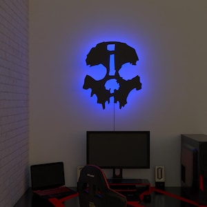 Puede incluir: Una lámpara de pared negra con forma de calavera con un resplandor de neón azul. La luz está montada en una pared blanca sobre una configuración de computadora. La habitación incluye un monitor de computadora, altavoces, una computadora portátil y una silla de juego.