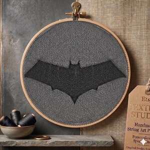 Pode incluir: Uma peça de arte em fio redonda, com o logotipo do Batman, emoldurada em um bastidor de bordado de madeira. O símbolo do morcego preto está centralizado em um fundo cinza. Uma pequena tigela de linha preta e um anel dourado estão em uma prateleira abaixo.