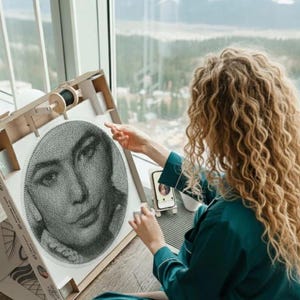 Könnte beinhalten: Eine Person arbeitet an einem Fadengemälde-Porträt eines Gesichts. Das Kunstwerk ist auf einem Holzrahmen mit einer Garnrolle befestigt. Die Person trägt ein tealfarbenes Oberteil und hat lockiges blondes Haar. Ein Smartphone ist sichtbar.