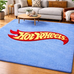 May include: A light blue rectangular rug with the Hot Wheels logo in red and yellow. The rug is placed on a hardwood floor in a living room setting. The logo is a stylized flame.