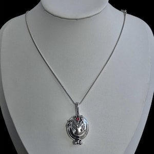 May include: A silver-toned metal locket pendant suspended on a thin silver chain. The oval-shaped locket features a raised, swirling decorative pattern and a single small, round, bright red gemstone set near the top of the pendant's front surface.