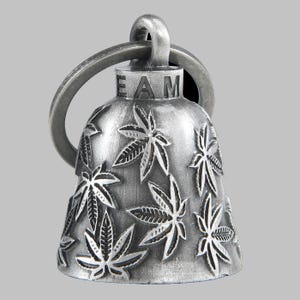 May include: A silver-colored bell with a ring at the top and a series of cannabis leaf designs. The word "EAM" is stamped on the top of the bell. The bell is likely made of metal and has a distressed finish.