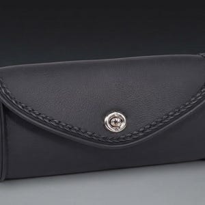 May include: A black rectangular leather pouch with a rounded flap closure. The flap features a braided leather trim along the edge and a central silver-toned metal twist-lock fastener. The pouch has a smooth, consistent texture throughout its surface.