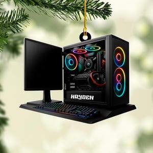 May include: A black computer ornament with a monitor, keyboard, and tower. The tower has colorful LED lights and the name "HAYDEN" on the front. The keyboard and lights have a rainbow color scheme. The ornament is hanging from a gold string.