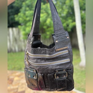 May include: A dark gray leather shoulder bag with multiple zippered compartments and pockets. The bag has a distressed appearance with gold-colored zippers and hardware. The bag is held up against a blurred green background.